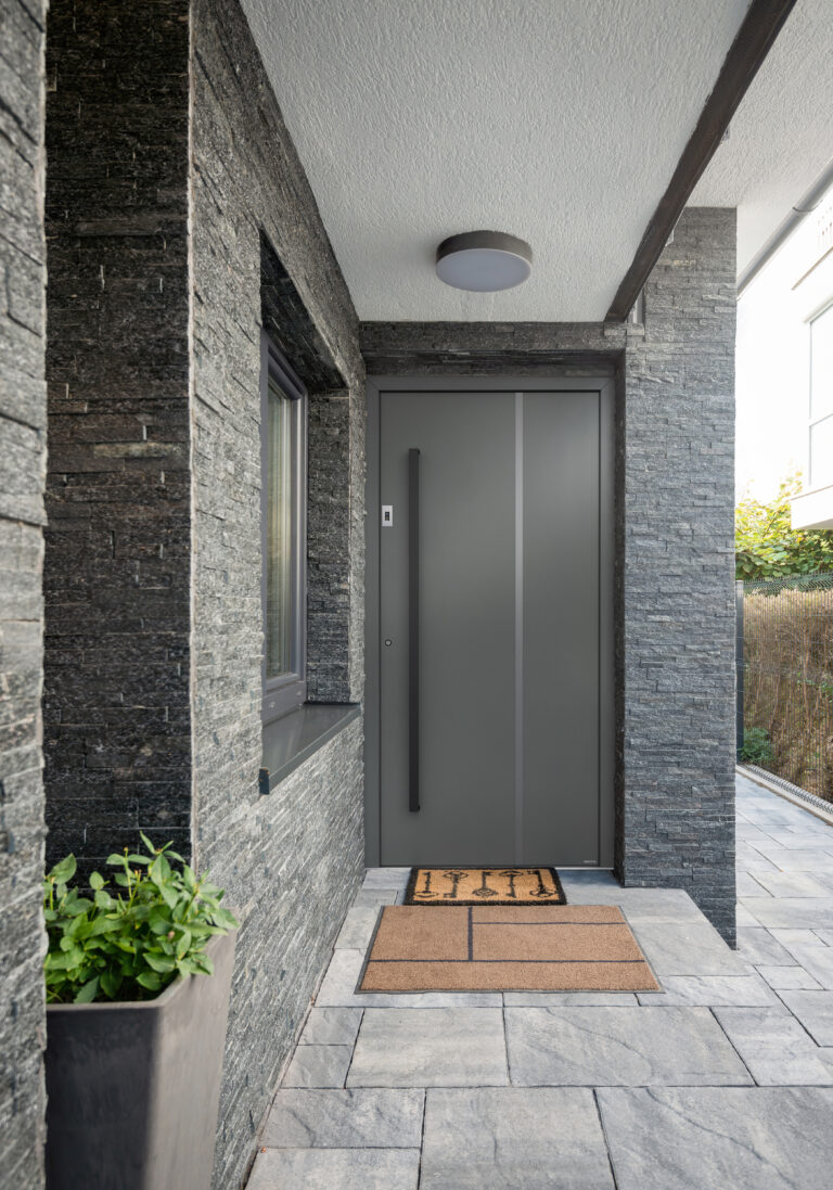 Das Bild zeigt einen modern gestalteten Eingangsbereich eines Hauses. Die Wände und Säulen sind mit dunkelgrauen, strukturierten Natursteinplatten verkleidet. Im Zentrum befindet sich eine große, matte Haustür in einem dezenten Grauton mit vertikalen Linien und einem langen, schwarzen Griff. Vor der Tür liegen zwei Fußmatten, eine davon mit Schlüsselmuster. Links befindet sich ein Fenster mit grauem Rahmen und auf der Fensterbank steht eine graue Pflanzschale mit grünem Gewächs. Die Bodenplatten sind großformatig und in einem hellen Grauton gehalten. Die Decke ist weiß und es ist eine runde, moderne Deckenleuchte installiert. Rechts sieht man einen Teil des Gartens mit lichtem Gebüsch. Der gesamte Bereich wirkt aufgeräumt, hochwertig und einladend.