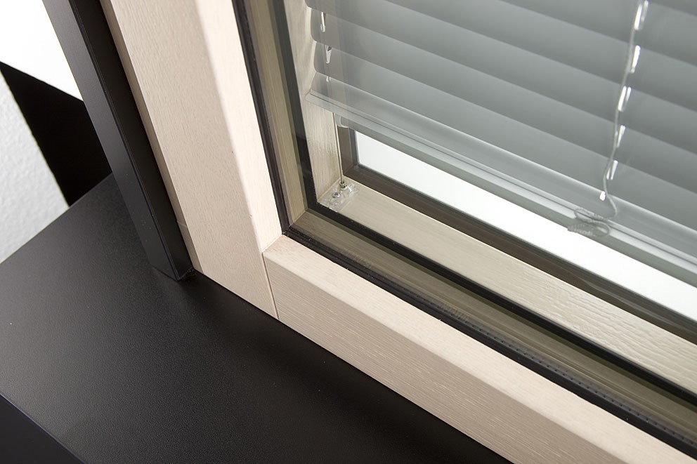 Das Bild zeigt eine Nahaufnahme eines modernen Fensterrahmens. Der Fensterrahmen besteht aus hellem Holz mit klaren, geraden Kanten. Die Verglasung ist doppelt ausgeführt, und im inneren Zwischenraum befinden sich horizontal verlaufende, silberfarbene Lamellen eines Jalousiesystems. Die Lamellen sind geschlossen und bieten Sichtschutz. Der Bereich um das Fenster ist dunkel gehalten, und die Aufnahme fokussiert auf die Ecke des Fensterrahmens sowie das Innenleben der Jalousie. Das Bild vermittelt hochwertige Verarbeitung und moderne Technik zum Thema Privatsphäre und Blendschutz. Fensterprofi in Neumünster und Umgebung