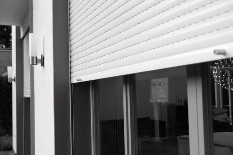 Detailansicht eines teilweise heruntergelassenen weißen Aluminium-Vorbaurollladens vor einer anthrazitgrauen Schiebetür-Verglasung an einer modernen Hausfassade, Schwarz-Weiß-Aufnahme.
