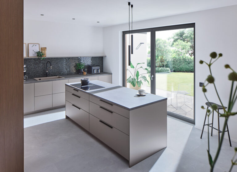 Moderne, helle Küche mit Kochinsel und zweiflügeliger Balkon- bzw. Terrassentür in dunklem Rahmen, durch die große Glasfläche fällt viel Tageslicht und der Blick auf den Garten. ​