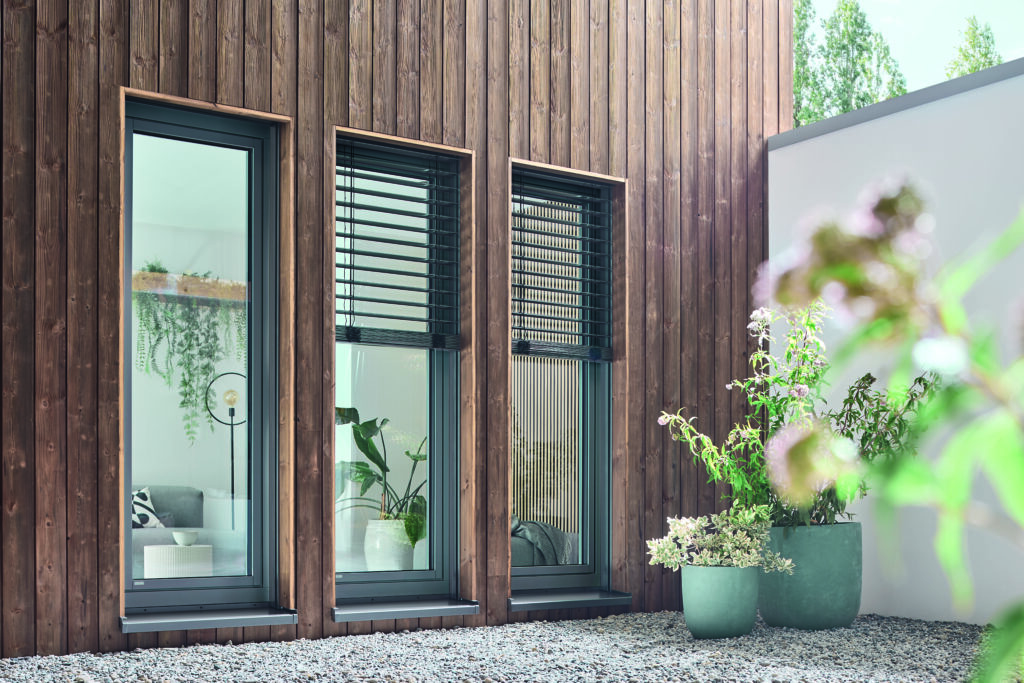 Das Bild zeigt die Außenansicht einer modernen Hausfassade mit senkrechter Holzverkleidung in warmen Brauntönen. Es sind drei große Fenster nebeneinander zu sehen, jeweils mit einem grauen Rahmen. Das mittlere und rechte Fenster sind mit Außenjalousien ausgestattet, die teils geöffnet und teils geschlossen sind. Im Inneren erkennt man eine helle und freundliche Einrichtung mit Pflanzen und einem Sofa. Vor den Fenstern auf dem Kiesbett stehen drei große Pflanzenkübel mit unterschiedlichen grünen und blühenden Pflanzen. Rechts daneben befindet sich eine helle Mauer, im Hintergrund sind hohe Bäume zu sehen. Das Gesamtbild vermittelt ein modernes und natürliches Wohngefühl