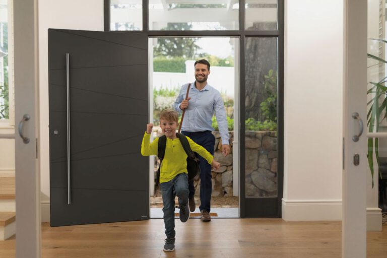 Eine moderne Eingangsszene: Ein Kind mit Rucksack läuft fröhlich durch eine geöffnete, große, dunkle Haustür in einen hellen Flur. Dahinter betritt ein Erwachsener, ebenfalls mit Tasche, das Haus. Im Hintergrund sind durch die Glastüren ein Garten und eine Steinmauer sichtbar. Die Stimmung wirkt freundlich und lebendig. Die Haustür hat ein schlichtes, elegantes Design mit einer langen senkrechten Griffstange und dezenten Linien