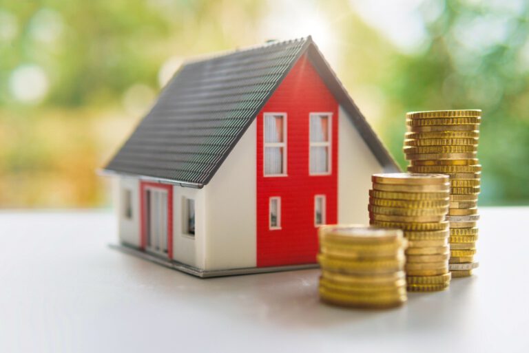 Miniaturhaus mit roter Fassade und grauem Satteldach neben drei ansteigenden Stapeln Goldmünzen auf weißer Fläche vor grün verschwommenem Hintergrund als Symbol für Fördermittel oder Immobilienfinanzierung.
