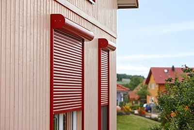 Zwei rote Vorbaurollläden mit gerundetem Rollladenkasten und geöffneten Lamellen an einer beigen Holzverkleidungsfassade, im Hintergrund eine grüne Wohnsiedlung.