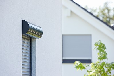 Nahaufnahme eines runden, edelstahlfarbenen Rollladenkastens eines Vorbaurollladens an einer weißen Hausfassade, im Hintergrund ein weiterer Rollladen am Nachbargebäude.
