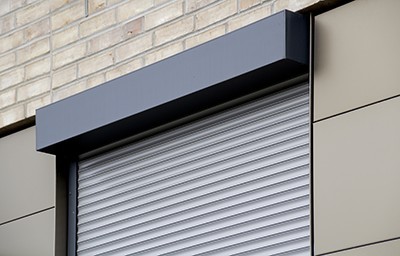 Nahaufnahme eines anthrazitgrauen Vorbaurollladens mit flachem, eckigem Rollladenkasten an einer gemischten Fassade aus Klinker und beigen Fassadenpaneelen.