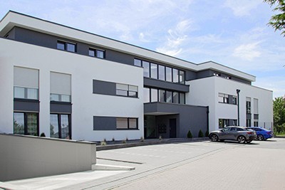 Modernes, zweigeschossiges Büro- oder Geschäftsgebäude in Weiß-Anthrazit mit zahlreichen Aufsatzrollläden vor großen Fensterfronten und gepflegtem Außenbereich.