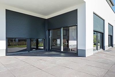 Eckansicht einer modernen Hausfassade mit anthrazitgrauen Aufsatzrollläden vor großen Schiebetüren und Fenstern auf einer hellgrauen Natursteintetrasse.